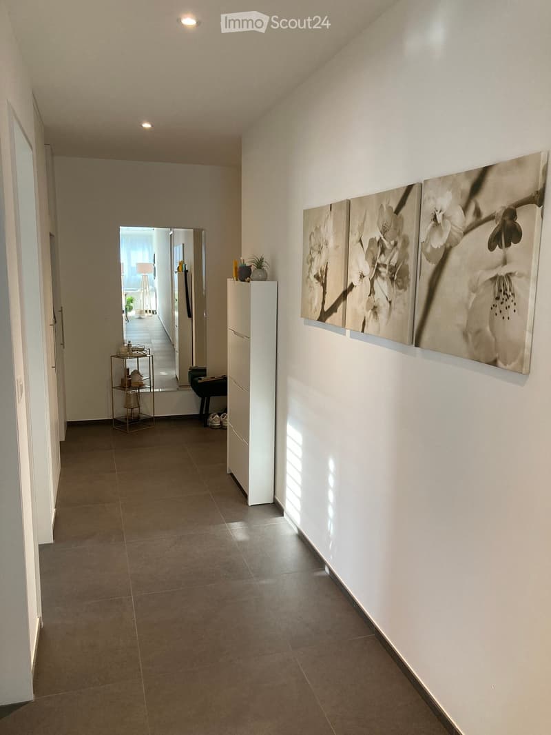 Modern hallway with tiled floor, white walls, recessed lighting, and artwork displayed on one wall. A small cabinet and a mirror are visible, with a glimpse of another room through an open doorway.