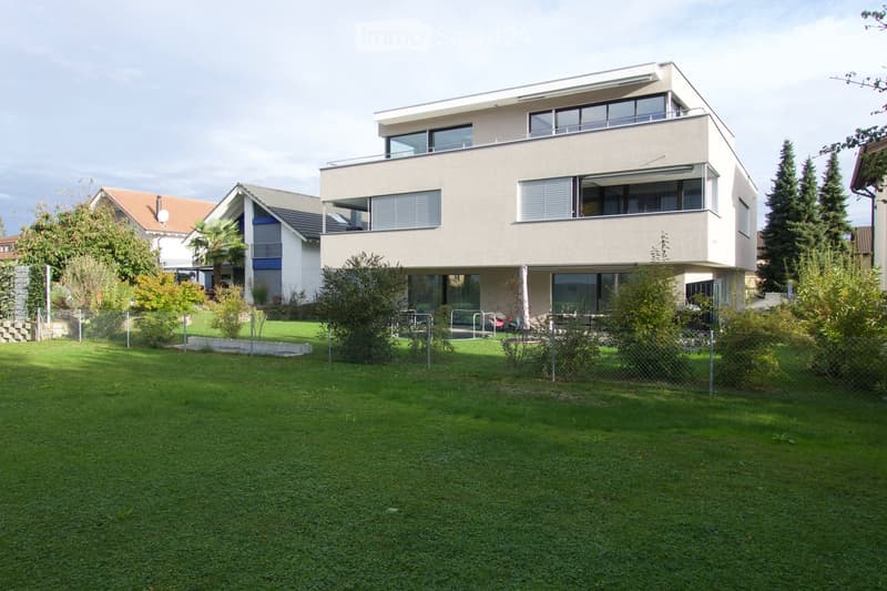 Modern two-story house with large windows and a balcony, surrounded by a well-maintained lawn and trees.