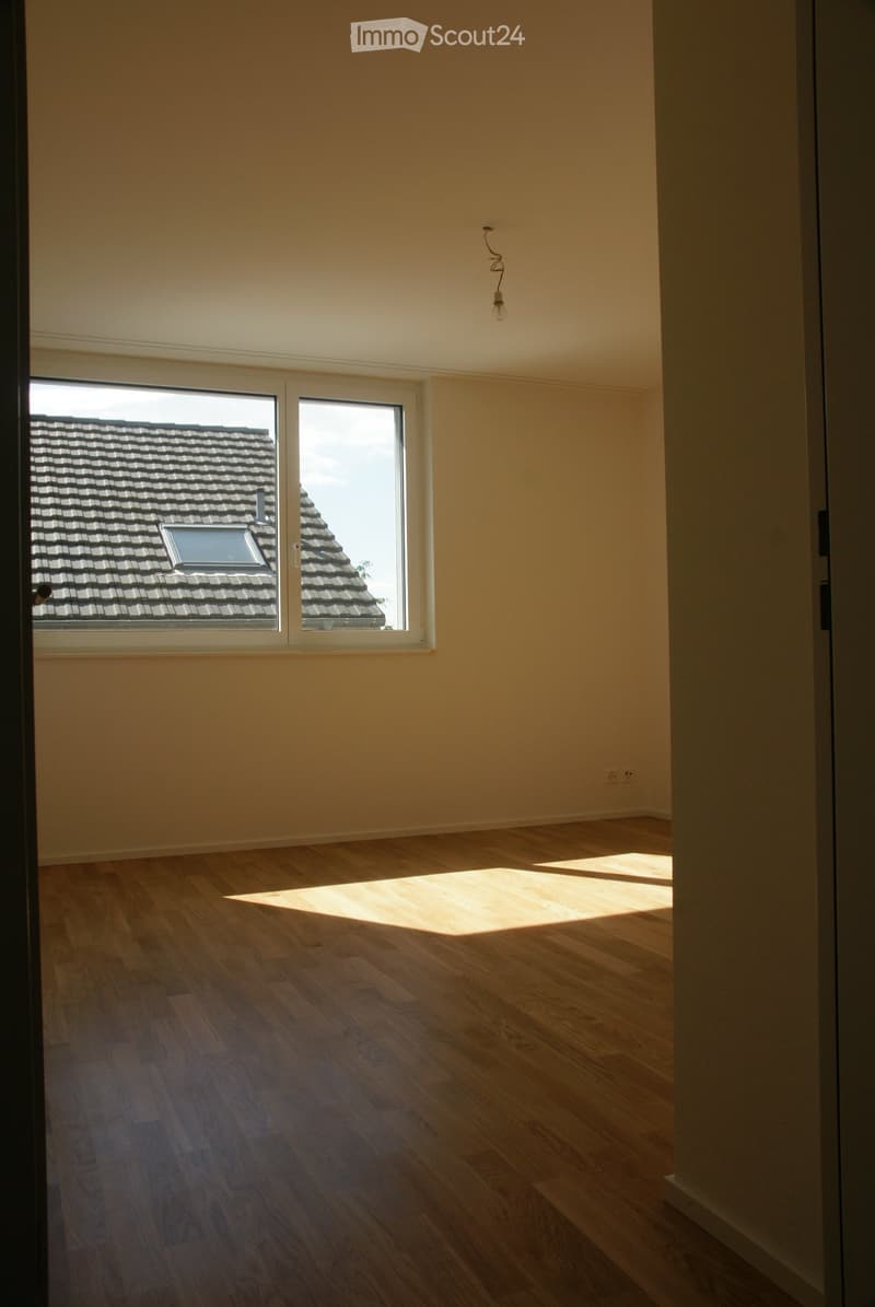 Empty room with light wooden flooring and large windows showing a roof view. No furniture or equipment present.