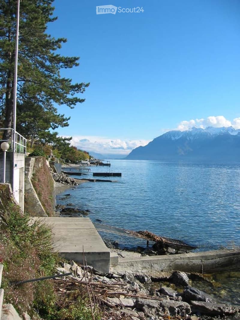 A scenic lakeside view with a concrete pier, calm water, distant mountains, and trees lining the shore.