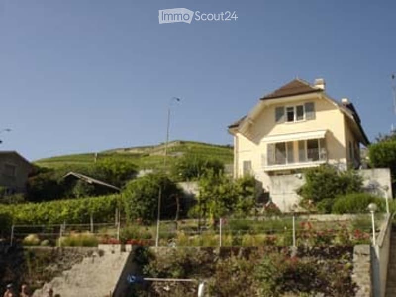 A two-story yellow house with white trim, situated on a hillside with a vineyard in the background. The house features a balcony and is surrounded by greenery and a stone retaining wall.