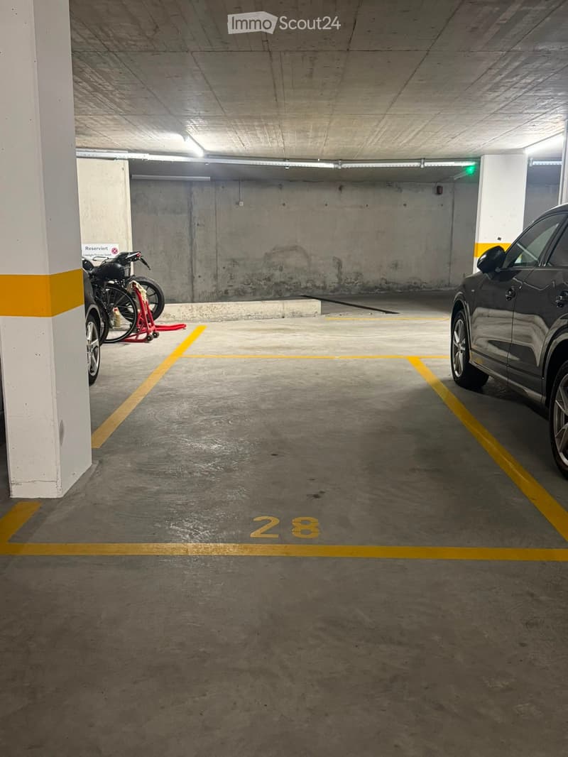 Underground parking area with concrete floors and walls, marked parking spaces, and a few vehicles including a car and a motorcycle.