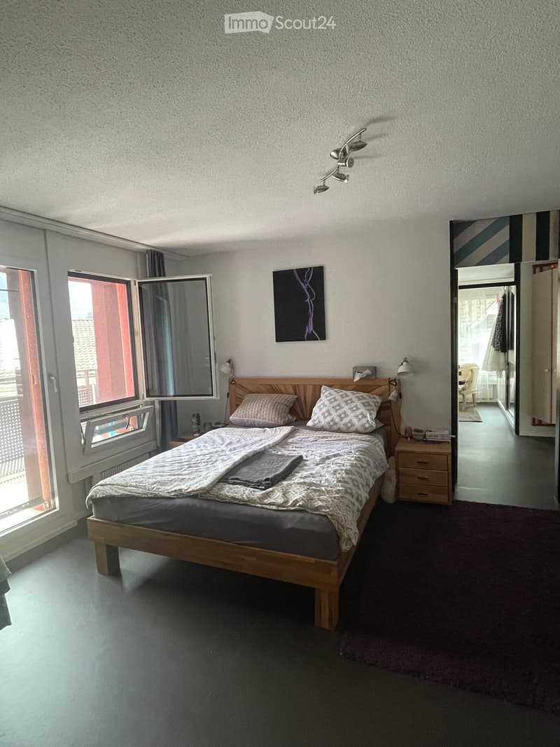 A well-lit bedroom with a wooden bed, nightstand, mirror, and artwork on the wall. The room features large windows with open blinds and a view of the outside.
