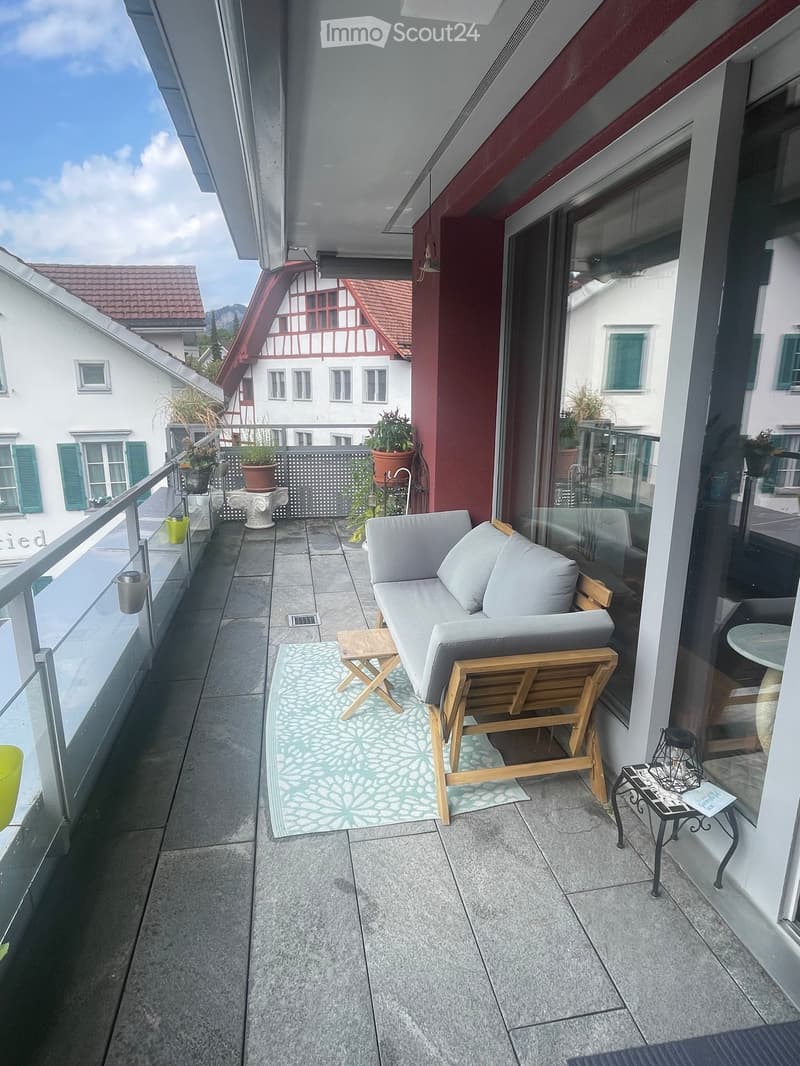 Balcony with grey sofa, wooden chair, small table, rug, and potted plants, overlooking a town with traditional houses.