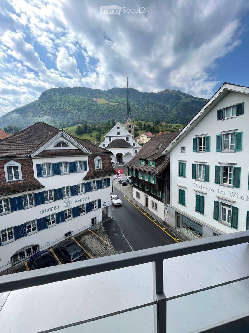 View from a balcony overlooking a street lined with traditional European buildings, including a hotel and a church with a tall spire, surrounded by green hills and mountains.