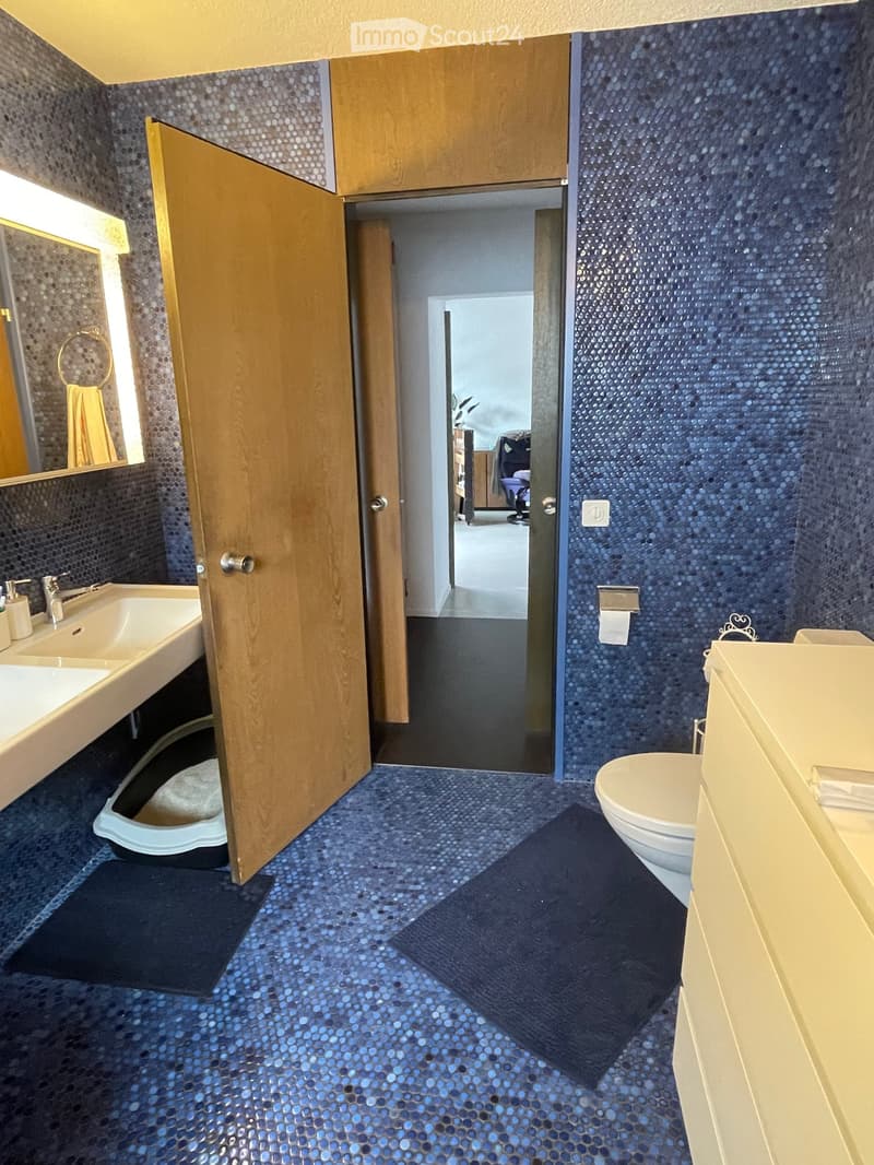 Bathroom with blue mosaic tiles, double sink, toilet, mirror, and wooden cabinet.