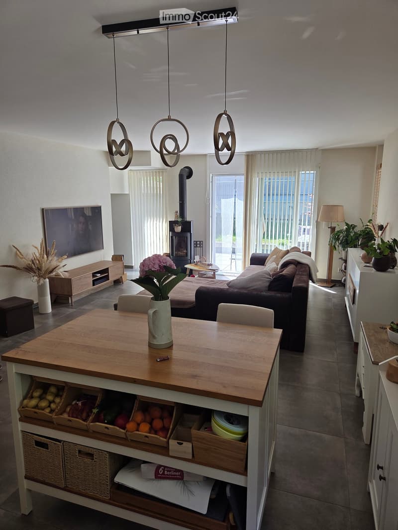 Modern living and dining area with a sofa, coffee table, fireplace, TV, dining table with chairs, pendant lights, and a kitchen island with storage baskets.