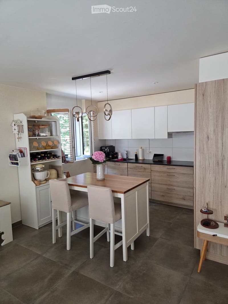 Modern kitchen with white cabinets, wooden countertops, and a central island table with two chairs. Features include a sink, coffee maker, laptop, vase with pink flowers, and a small shelf with jars and a mixer.