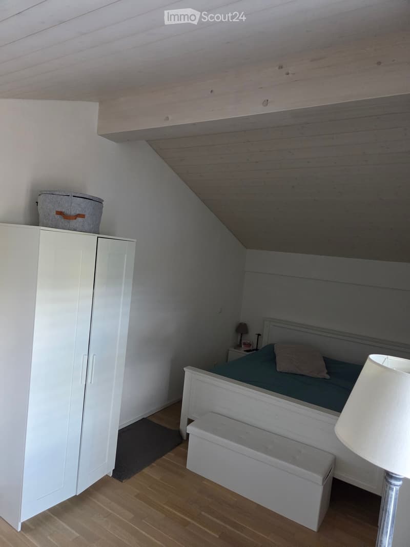 Cozy attic bedroom with wooden slanted ceiling, white wardrobe, grey bed with blue bedding, nightstand with lamp, and a small bench at the foot of the bed.