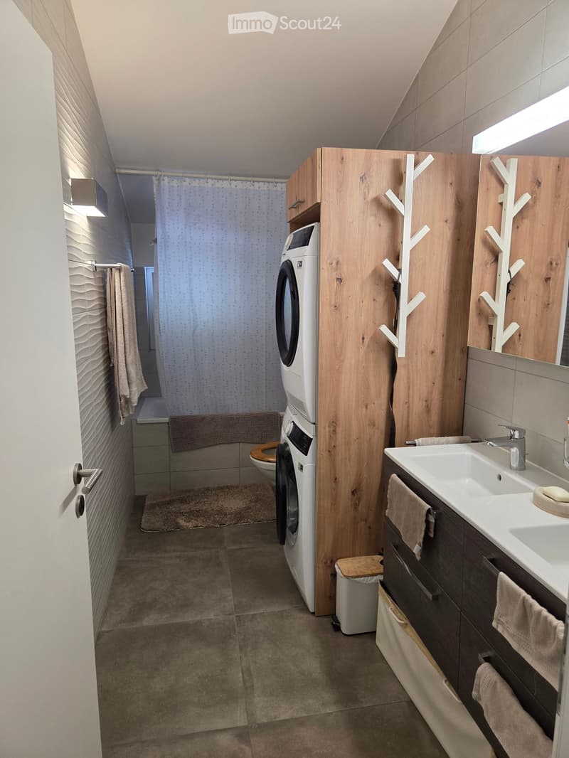 Modern bathroom with a sink, toilet, washing machine, and shower area. Features wooden cabinets, a white sink, and a shower curtain.