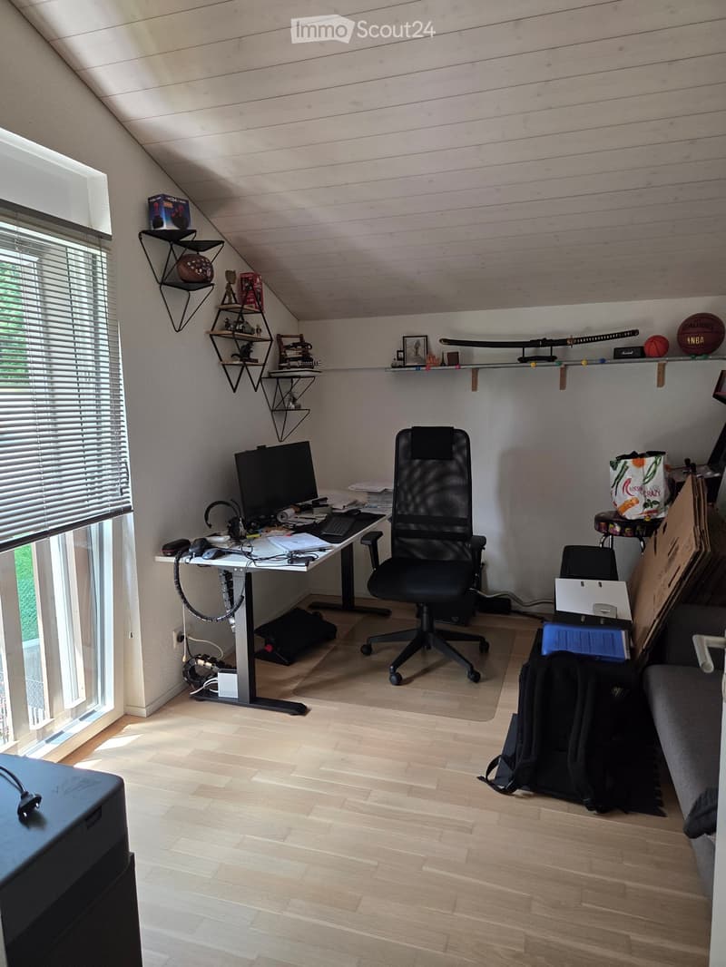 A modern, well-lit attic workspace with a standing desk, ergonomic chair, computer setup, and decorative shelves holding sports memorabilia and books.