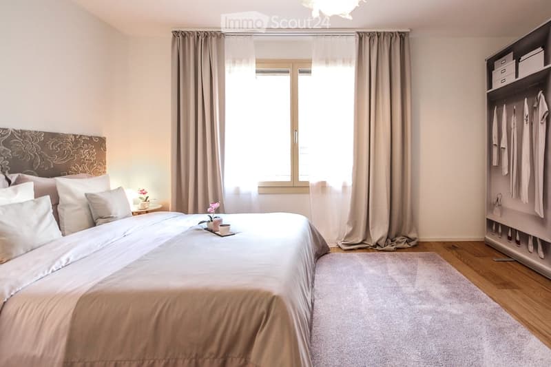 A bedroom with a bed, pillows, and a nightstand with a flower vase. A window with curtains and a lamp. A closet with clothes hanging on the racks.