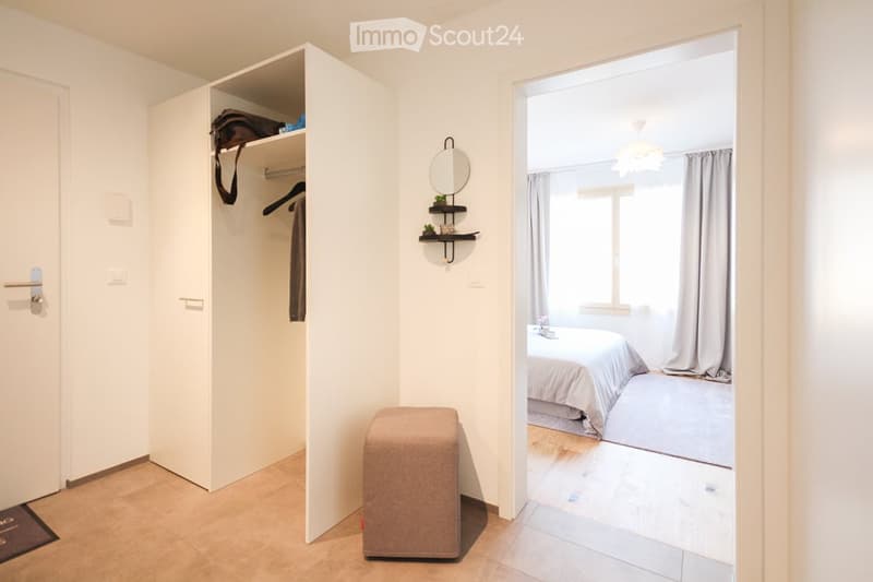A room with a white wardrobe, a mirror, a shelf, a stool, and a bed in the adjacent room.
