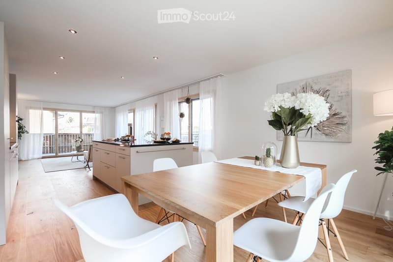 Dining room with wooden table, white chairs, white walls, wooden floor, kitchen island, white curtains, painting, flowers, plants