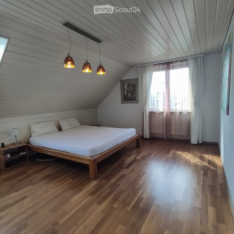 Bright bedroom with wooden floors, slanted ceiling, large window with curtains, bed with white linens, nightstand with lamp, and artwork on the wall.