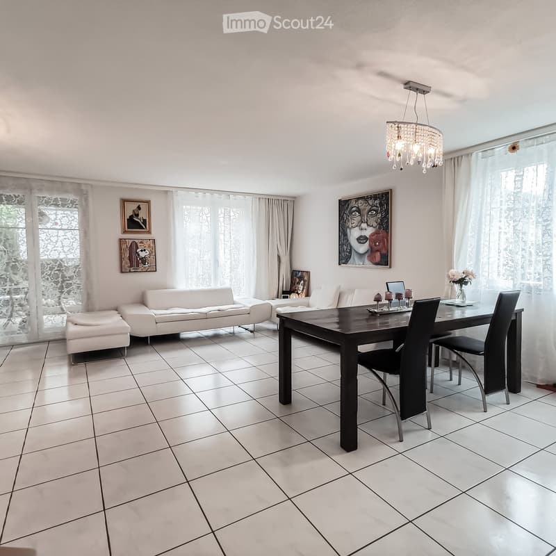 Bright living and dining area with white walls, tiled floor, large windows with curtains, a white sofa with ottoman, a dark wooden dining table with chairs, and a chandelier.