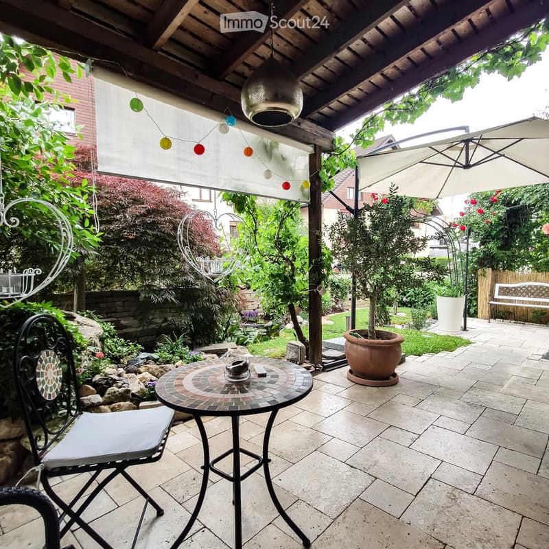 A covered outdoor patio with a round mosaic table, a folding chair, a large umbrella, a bench, and potted plants.