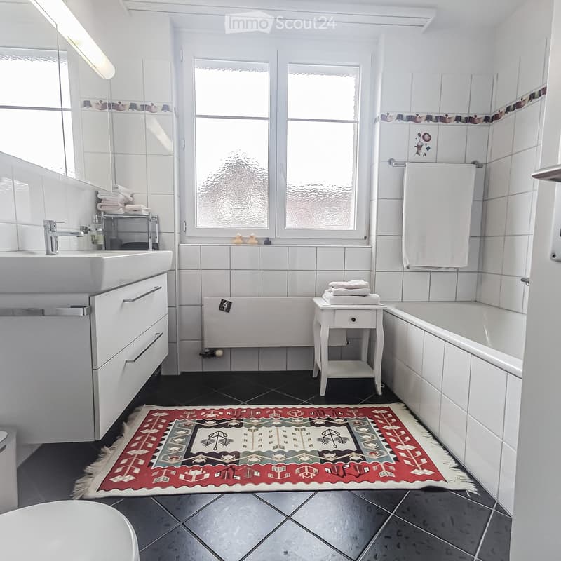 Modern bathroom with white tiles, a large window, a sink with a mirror cabinet, a bathtub, a small table with folded towels, and a decorative rug on the floor.