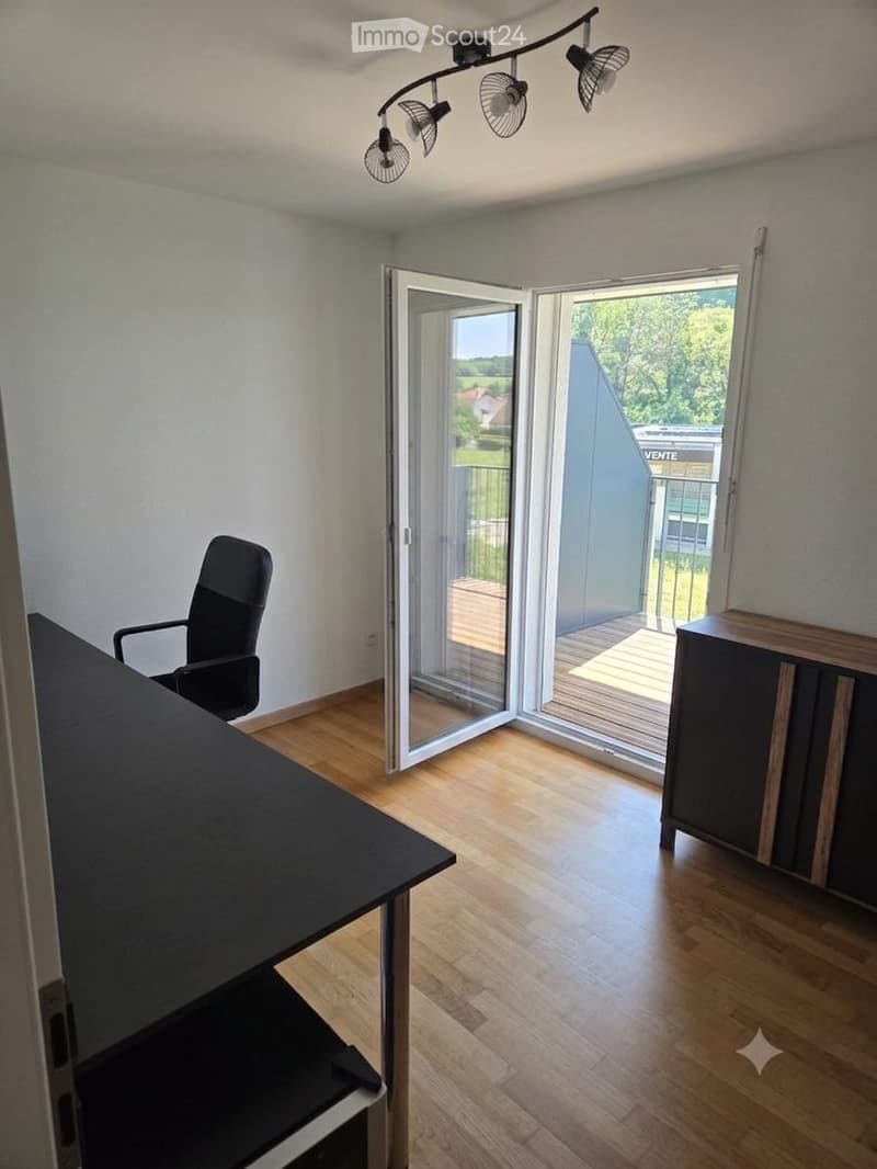 Bright room with wooden flooring, large sliding glass doors leading to a balcony, a black office chair, a black desk, and a dark wooden cabinet.