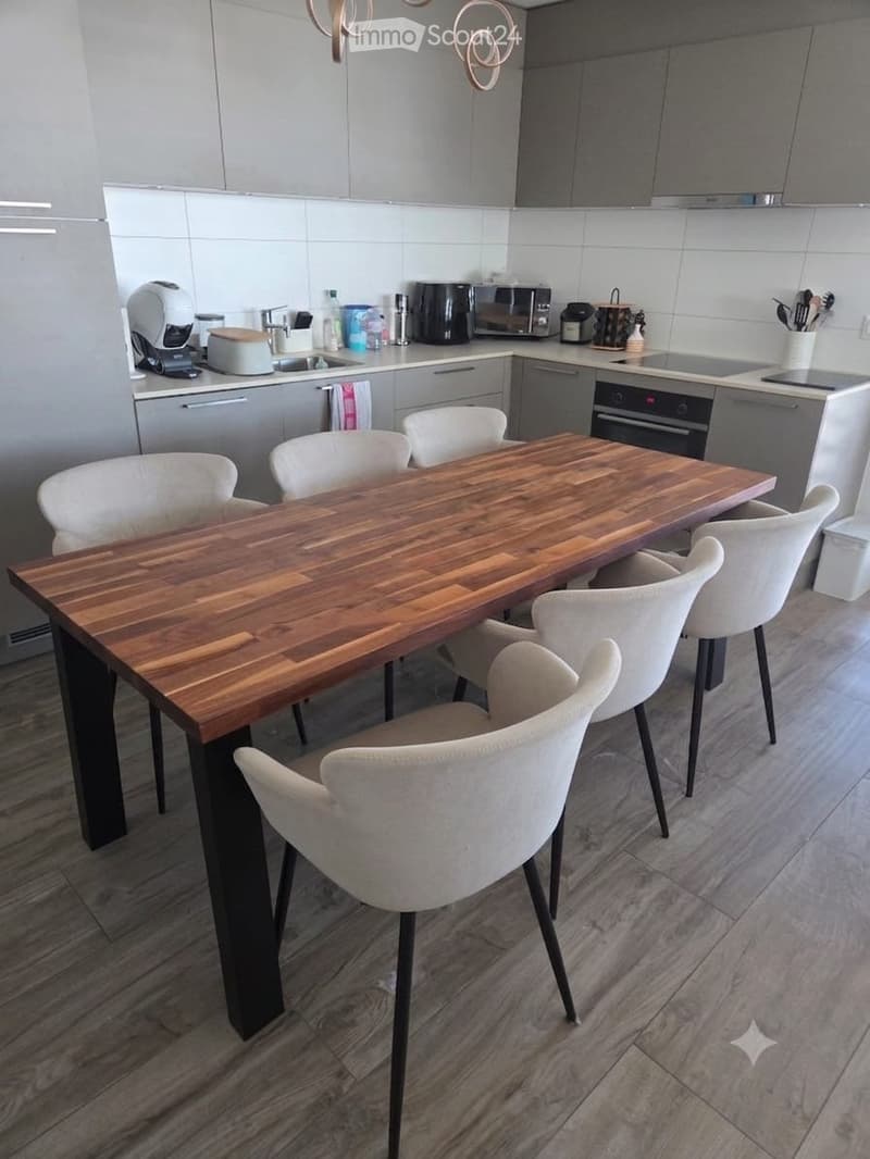 Modern kitchen with grey cabinets, a wooden dining table, white chairs, and various appliances including a coffee machine, oven, and microwave.