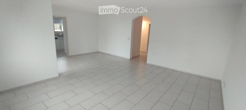 Empty room with white tiled flooring and white walls, featuring an arched doorway leading to another room and a window with blinds.