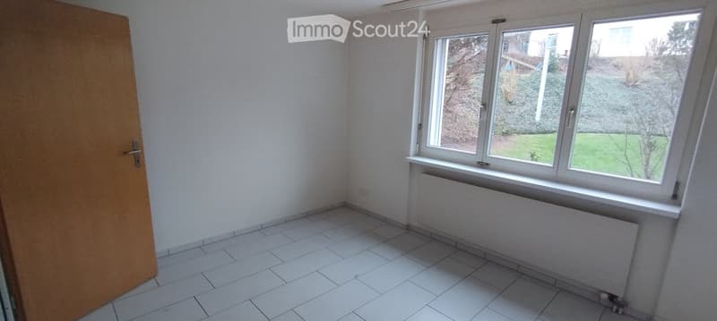 Empty room with white tiled floor, white walls, a wooden door, and a large window with a view of a garden.