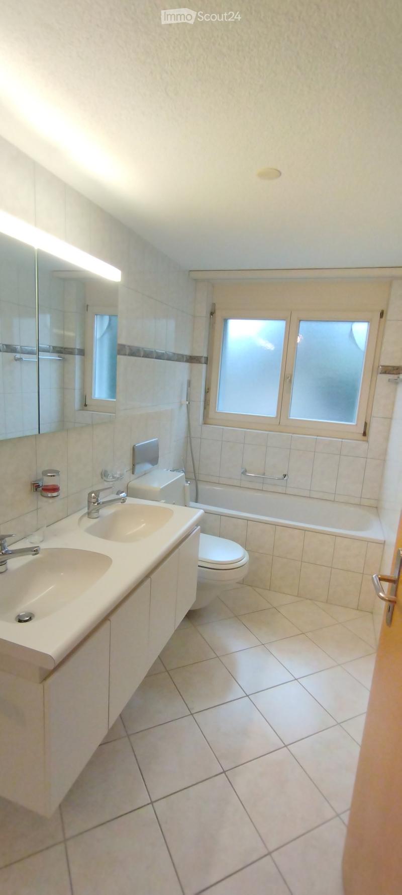 Modern bathroom with white tiles, double sink, toilet, bathtub, and large mirror. Features a window with frosted glass panels.