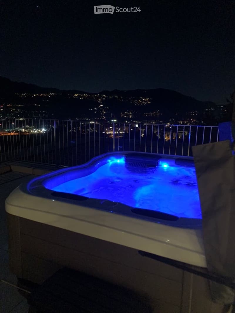 A rooftop terrace at night with a hot tub illuminated by blue lighting, overlooking a cityscape with lights and distant mountains.