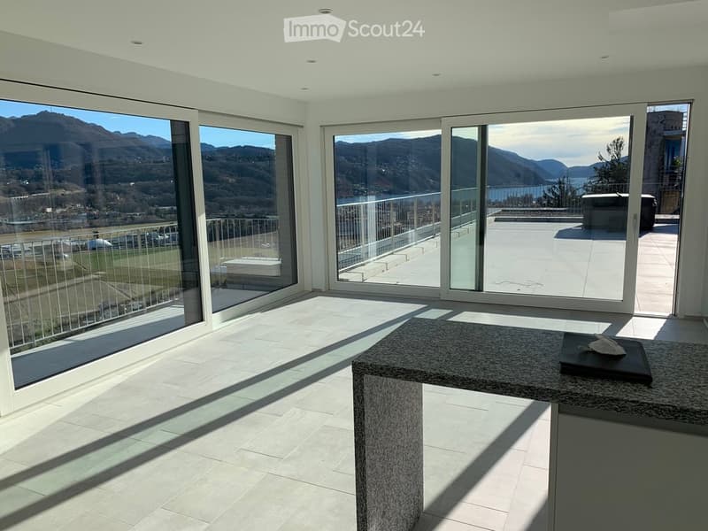 Bright, empty room with large glass sliding doors leading to a balcony, offering scenic mountain and lake views. The room features a tiled floor and a small granite countertop with a black tray on it.