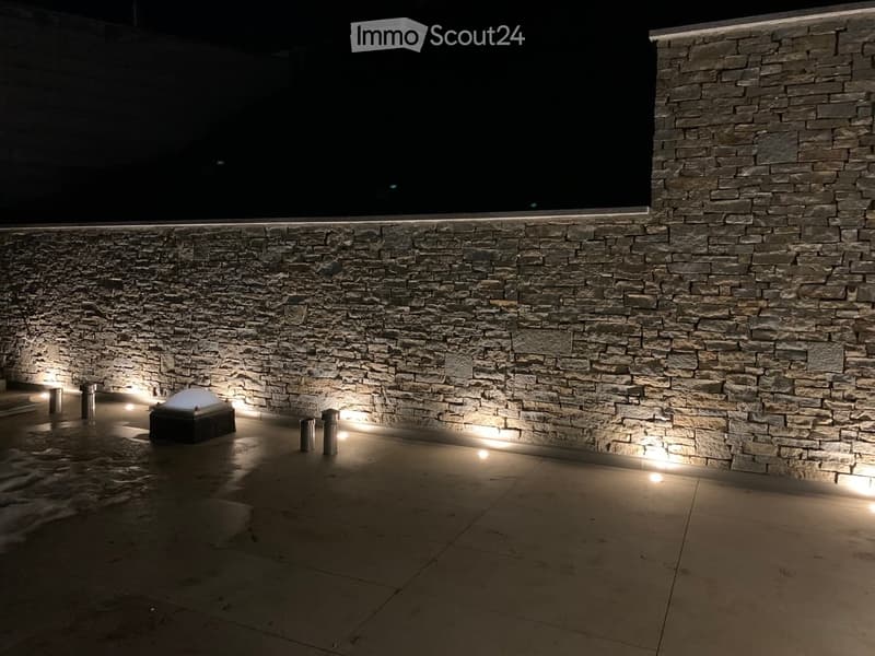 Nighttime view of a modern outdoor area with a stone wall, illuminated by recessed lighting, featuring a small black structure and cylindrical posts.