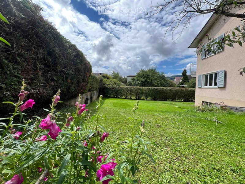 Well-maintained garden with green grass, flowering plants, and a hedge border, adjacent to a house with shutters and a window.