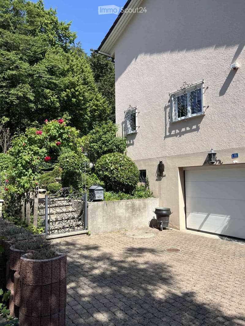Two-story house with beige exterior, garage, decorative wrought-iron window grilles, and a landscaped garden area with a gate and potted plants.