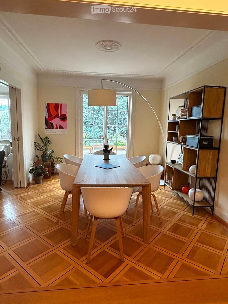 Dining room with wooden herringbone floor, long wooden dining table surrounded by white chairs, modern shelving unit with decorative items, and large windows with view of greenery.