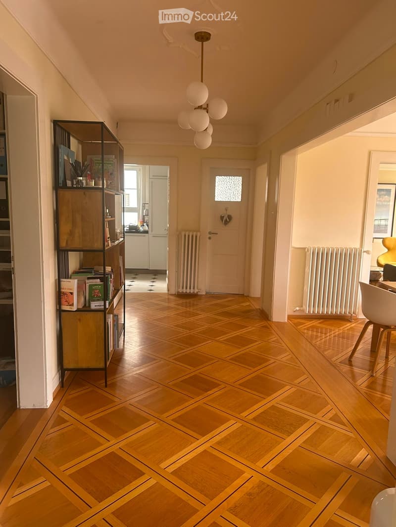 Bright hallway with wooden herringbone flooring, white walls, and a modern chandelier with spherical light fixtures. A black shelving unit with books and decorative items is visible on the left, and a white dining chair is seen on the right.