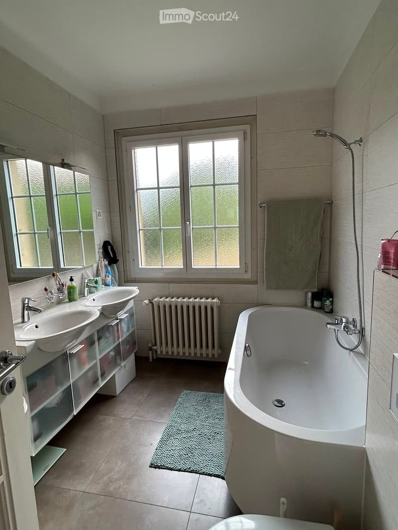 Modern bathroom with a freestanding bathtub, double sink vanity, large windows, and a radiator. Includes a bathroom rug and various toiletries.