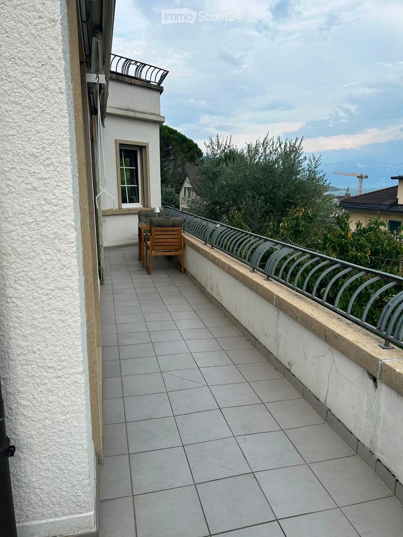 Balcony with tiled floor, metal railing, wooden chair, small table, and scenic view of greenery and distant landscape.
