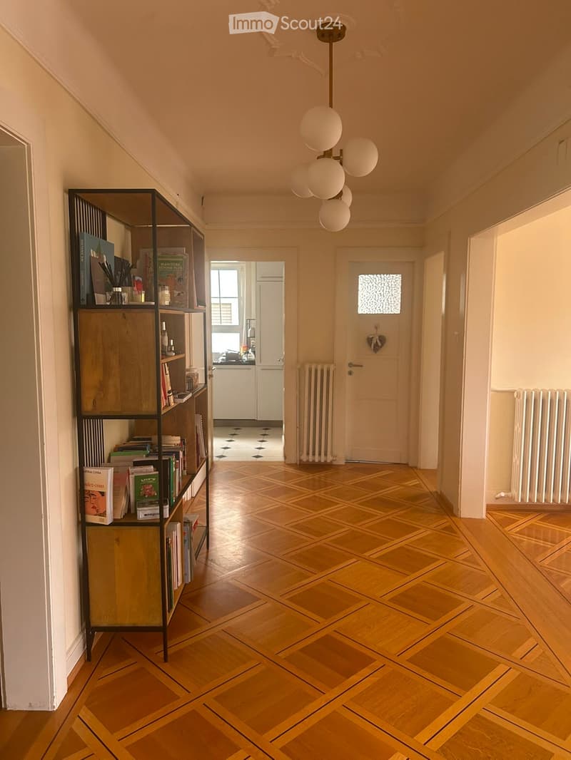 Hallway with wooden parquet flooring, white walls, a modern chandelier with spherical lights, and a black shelving unit with books and decorative items. Radiators are visible on both sides of the hallway.