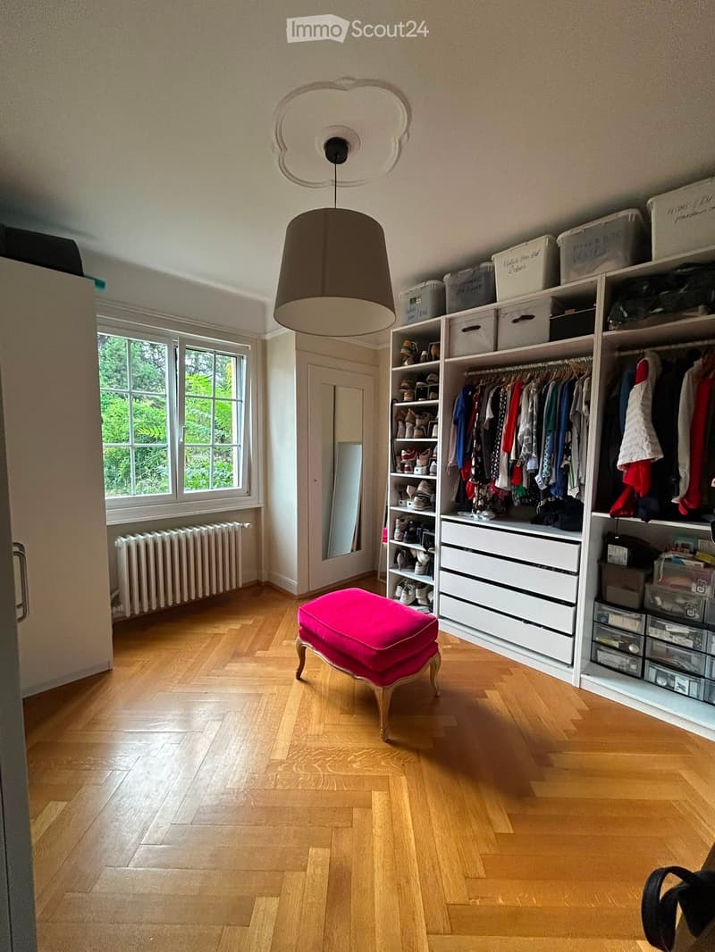 Well-lit room with wooden flooring, large window, built-in wardrobe with hanging clothes and shelves, storage boxes, a pink stool, and a mirror.