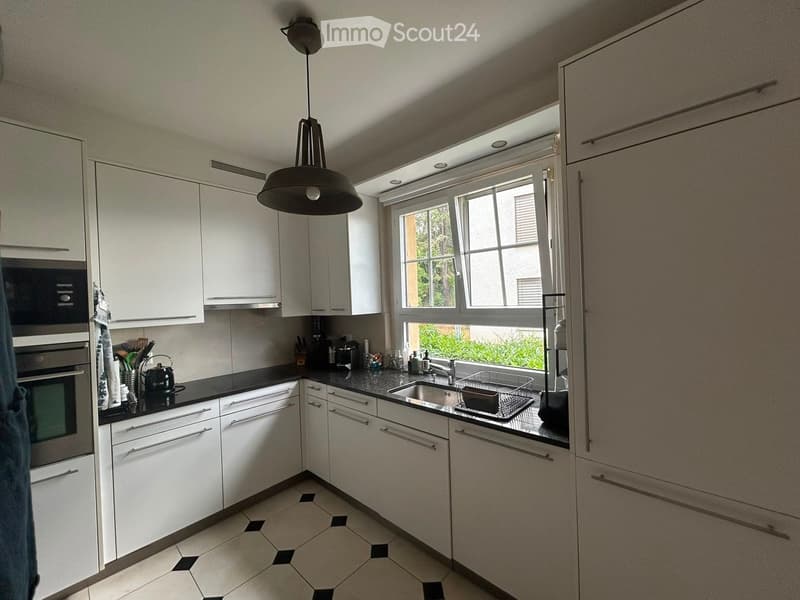 Modern kitchen with white cabinets, black countertops, and a large window. Features an oven, sink, and various kitchen utensils.