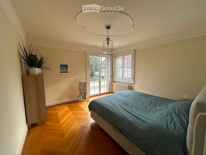 Bedroom with wooden floor, large window, bed with blue duvet, wardrobe, plant, picture frame, radiator, and a hanger.