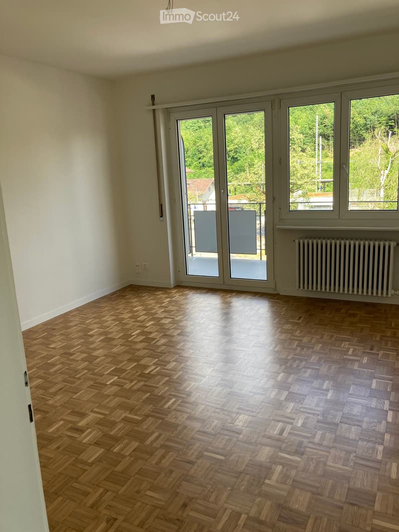 Empty room with wooden herringbone flooring, large windows, and sliding glass doors leading to a balcony.