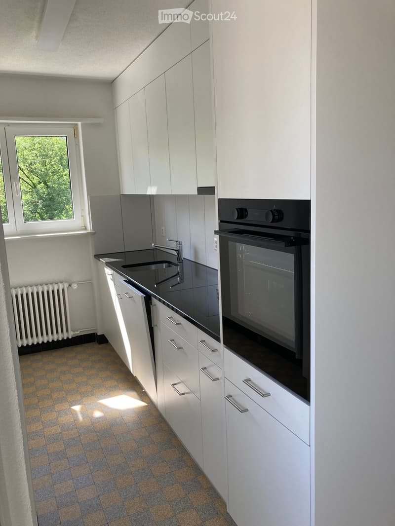 Modern kitchen with white cabinets, black countertop, stainless steel sink, and built-in oven. Features a checkered tile floor and a window with a view of greenery.