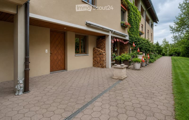 A well-maintained garden path leads to a house entrance, featuring neatly stacked firewood, potted plants, and decorative elements like a small statue and Swiss flags.