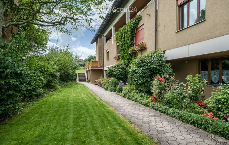 Well-maintained garden with a neatly trimmed lawn, a paved pathway, and a modern house with balconies and climbing plants.