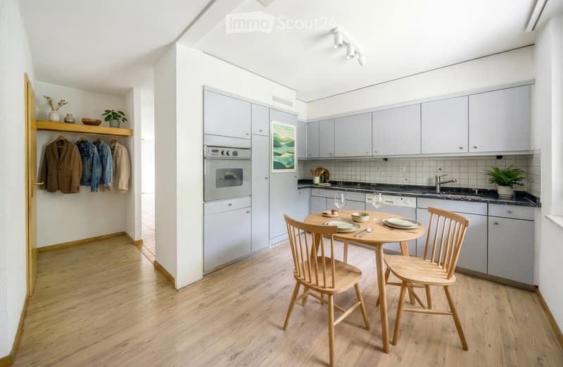 Modern kitchen with light grey cabinets, stainless steel appliances, a small dining table with two chairs, and a coat rack with jackets and a plant on the shelf.