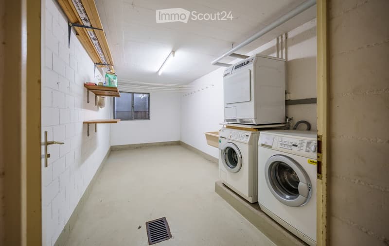 A small, well-lit laundry room with white tiled walls, a washing machine, a dryer, and wooden shelves on the left side.