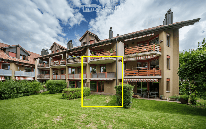 Multi-story residential building with balconies, awnings, and a well-maintained lawn in front.