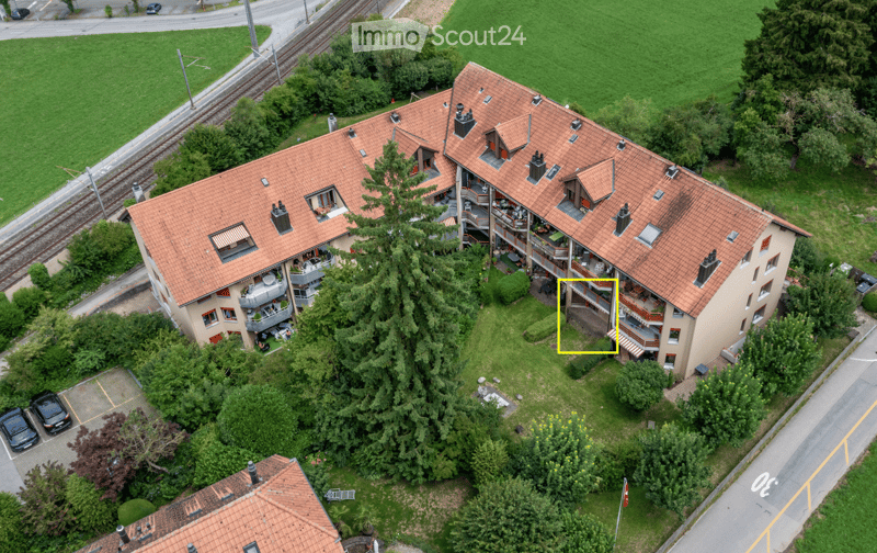 Aerial view of a multi-story residential building with red-tiled roofs, balconies, and surrounding greenery. The building has multiple apartments with visible outdoor seating areas and railings. There is a parking area with cars and a nearby railway track