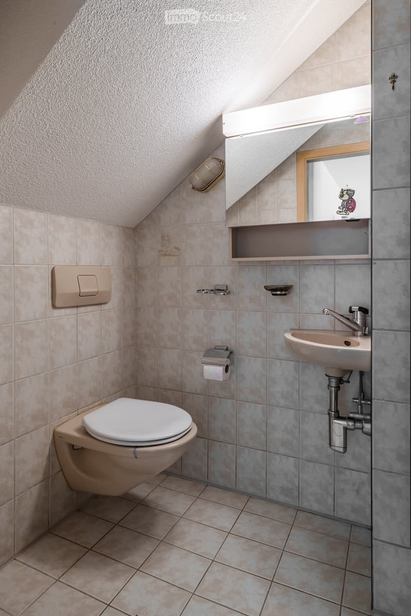A small, well-lit bathroom with beige tiles, a toilet, a sink with a mirror above it, and a toilet paper holder on the wall.