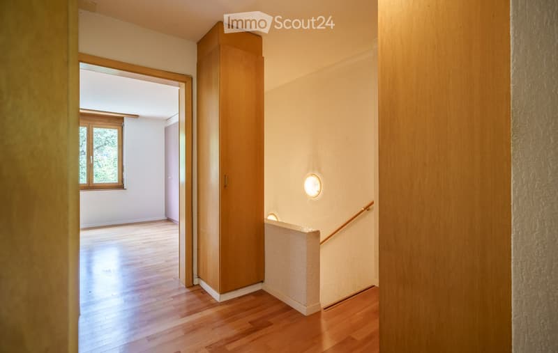 Empty hallway with wooden flooring, white walls, a built-in wooden cabinet, and a staircase leading upwards. A window at the end allows natural light to enter.